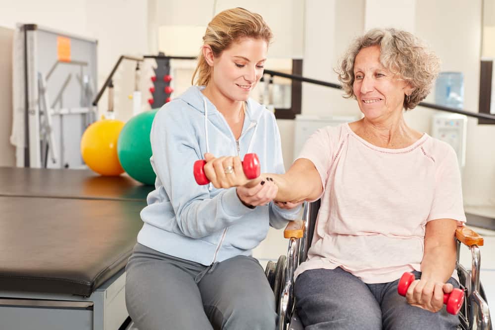 A physical therapist helps a senior woman using a wheelchair exercise her arms